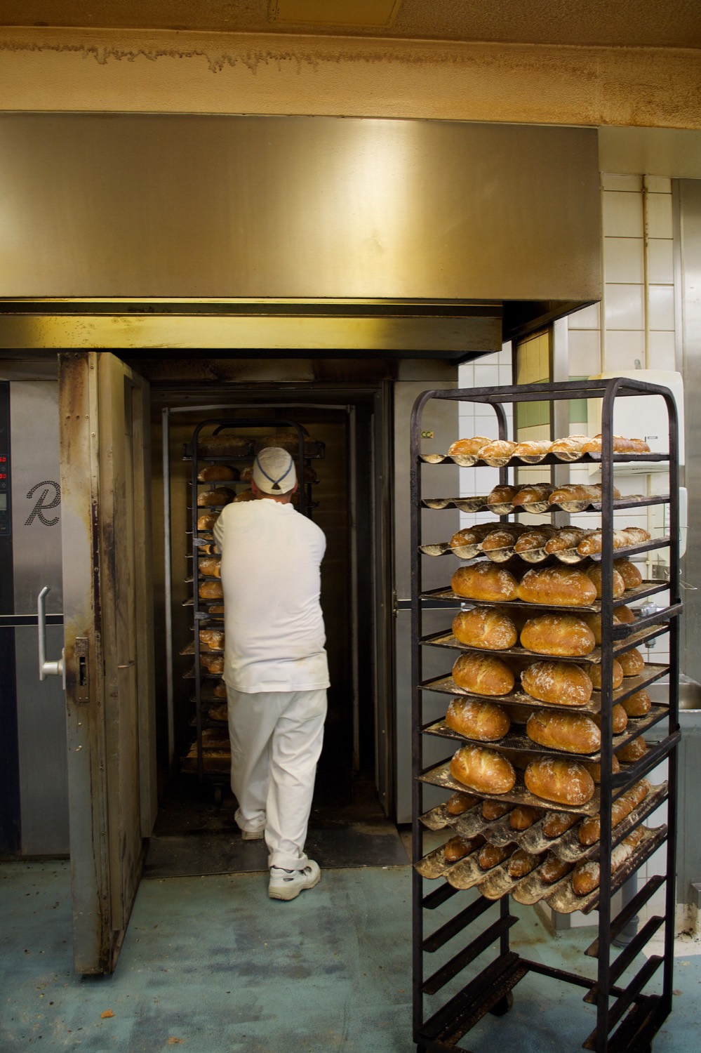 A baker in white wheels a rack of bread into a large rack oven, next to a second rack full of baked loaves.