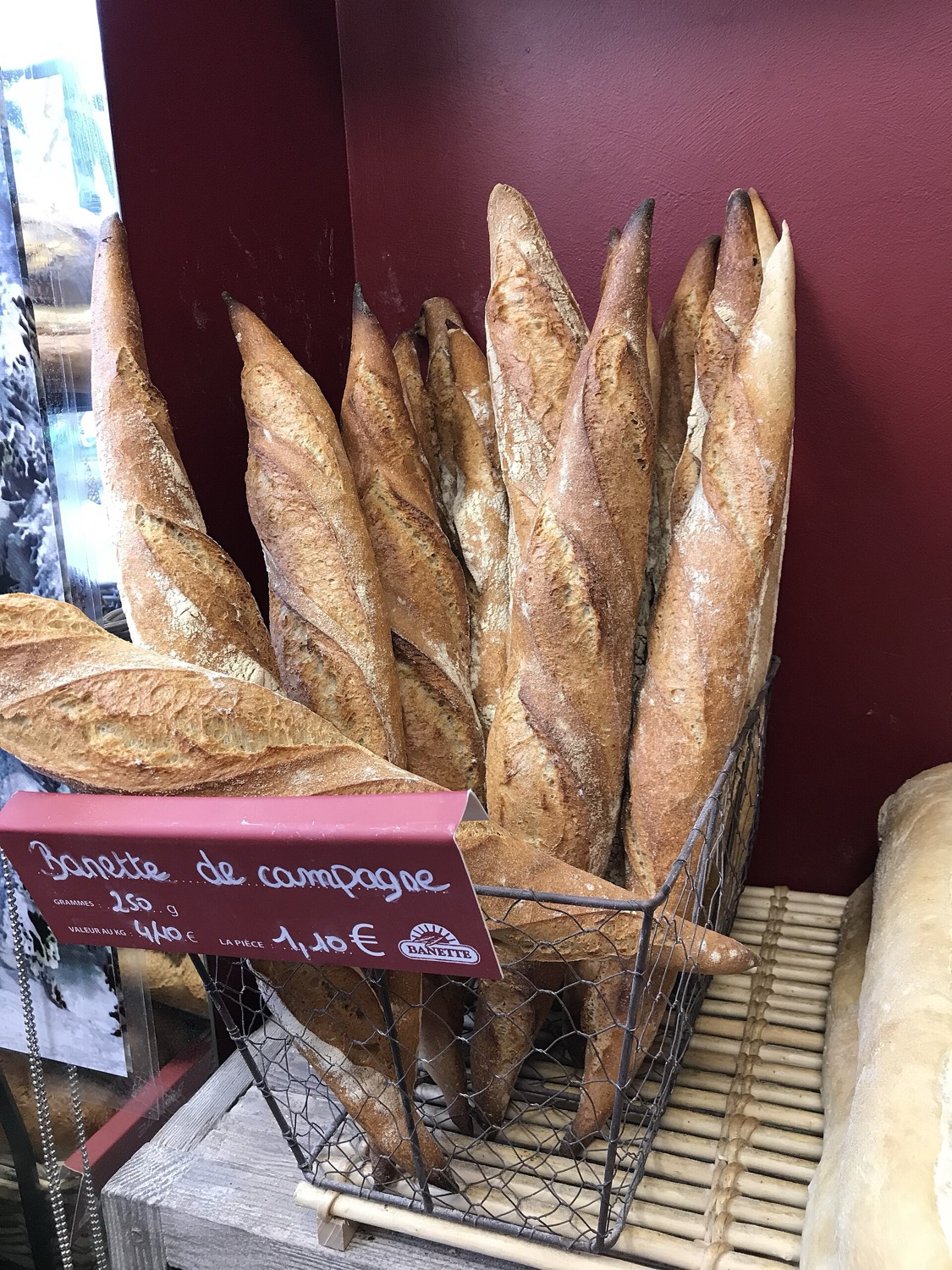 A wire basket of long rustic baguettes in a French bakery, with a red price card reading Baguette de campagne, 250 g, 1,10 €.