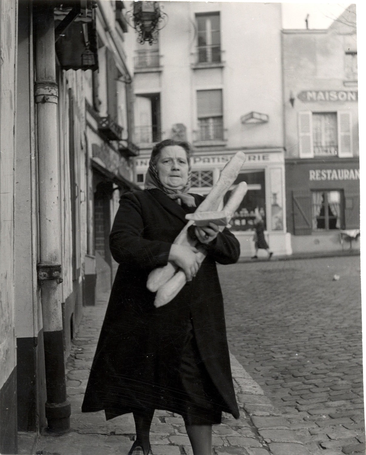 Black and white photograph: a woman in a dark coat walks down a cobbled Paris street with two baguettes under her arm.